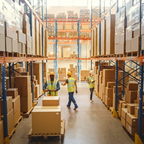 Three warehouse workers hard at work moving boxes up and down an aisle.